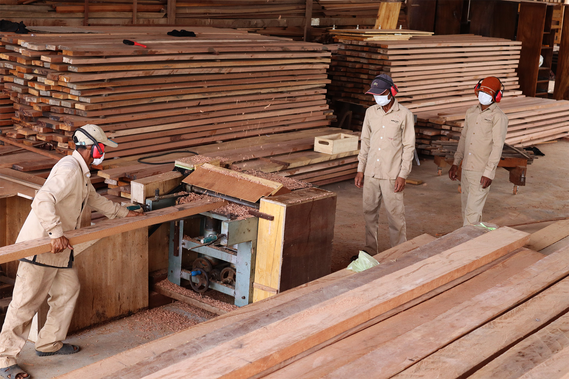 Des employés se tiennent entre les machines et les piles de bois de la menuiserie.