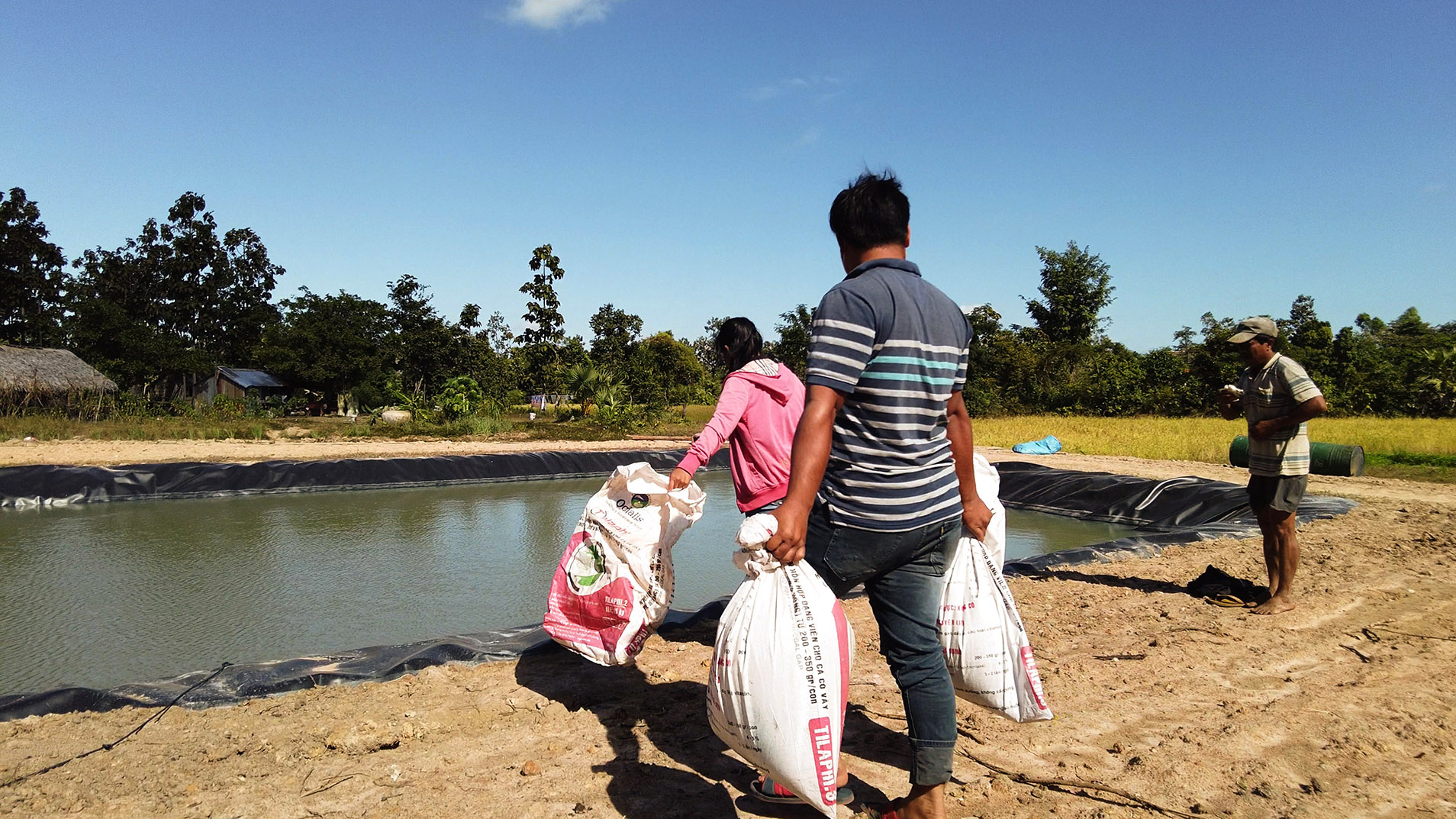 People carrying sacks beside a fish pond.