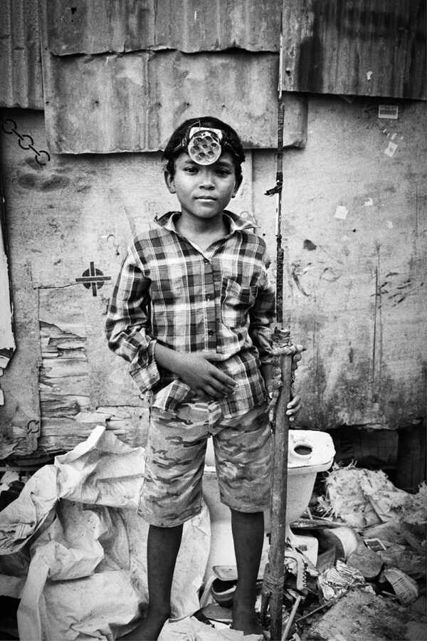 Portrait en noir et blanc d’un garçon devant une habitation modeste.